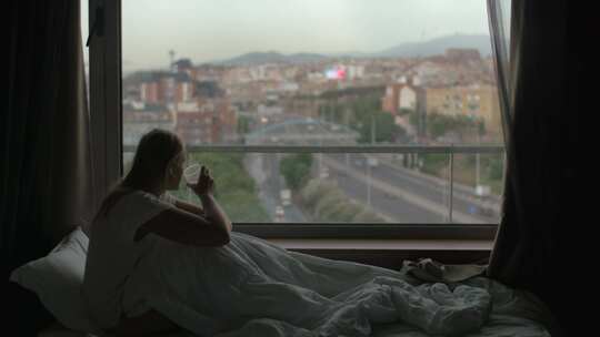 女人在床上喝咖啡，透过窗户看城市