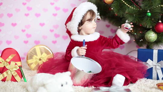 穿着红裙子的小女孩坐在圣诞树旁玩蜡笔