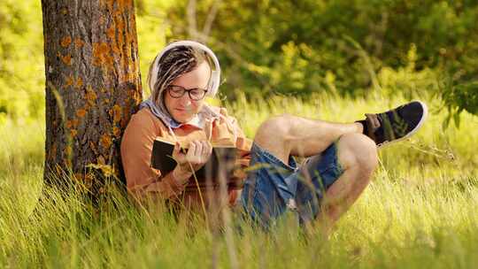 戴着眼镜，戴着耳机看书的年轻人，夏天坐在树旁。