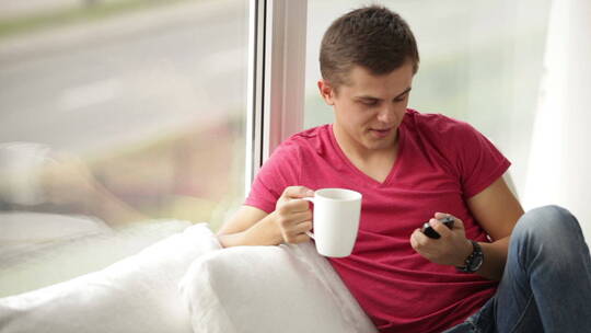 年轻人坐在窗边端着一杯茶看着手机傻笑