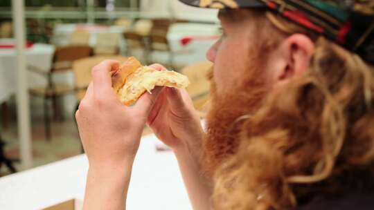 饥饿的红胡子男人吃奶酪比萨饼，坐在街头咖