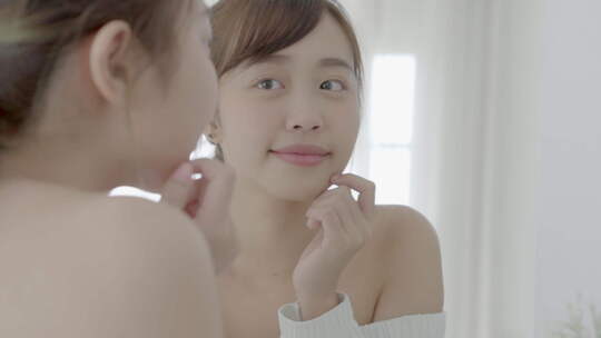 美丽的年轻亚洲女人微笑着看着健康检查皮肤护理高加索人的镜子。