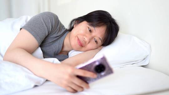 周末早晨年轻都市女孩懒在床上玩手机视频素材模板下载