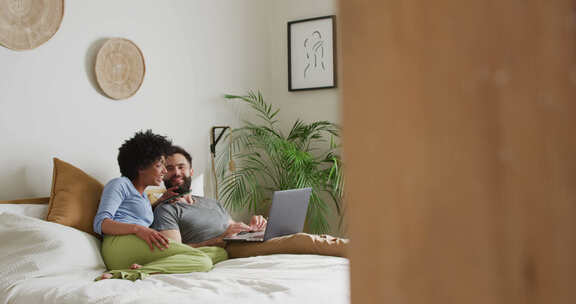 快乐的多样化夫妇在家放松的视频，躺在床上使用笔记本电脑和智能手机，复制空间