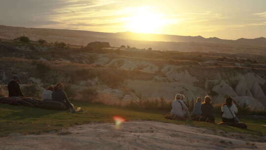人们坐在山上草地的垫子上，看着温暖的日落