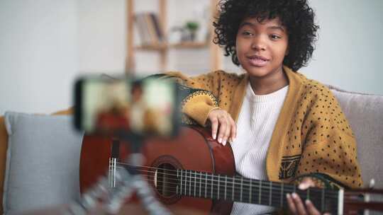 美丽的美国年轻女子将手机放在三脚架上并录制视频封面弹吉他和唱歌