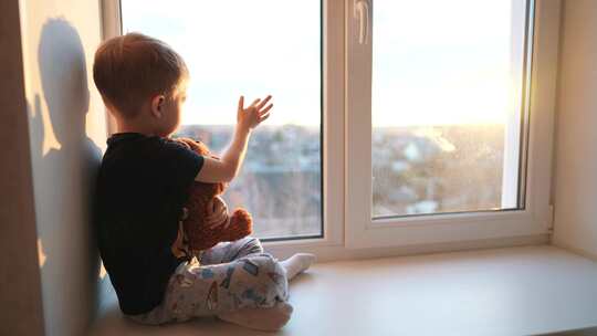 小男孩抱着小熊坐在窗户上，用手触摸阳光