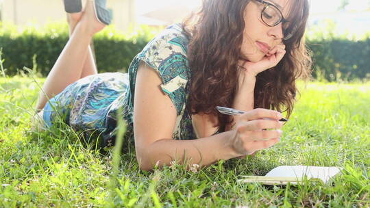 女人爬在草地上在记事本上写字视频素材模板下载