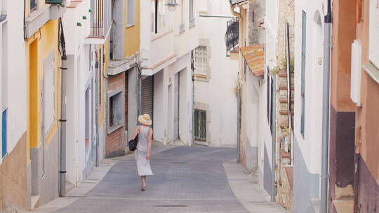 穿着浅色连衣裙戴着帽子的女游客走在老城的狭窄街道上