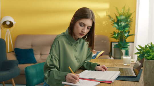年轻漂亮的女学生坐在桌旁做笔记在家学习