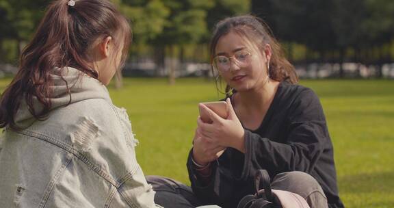 两位女大学生坐在校园草地上看手机聊天