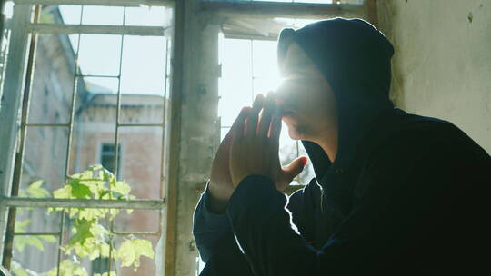 戴着兜帽的青少年坐在窗台上双手放在头上祈祷