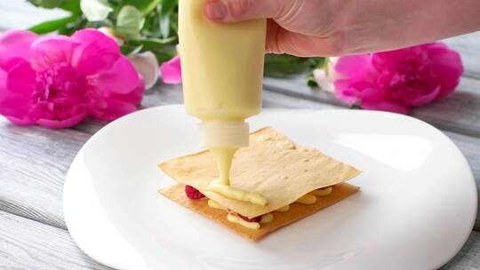 手工把奶油放在酥饼上。盘子旁边放着花和糕点。厨师准备覆盆子米莱菲耶。浆果分层甜点。