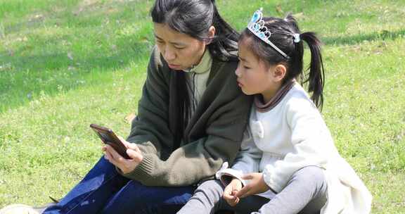妈妈带女儿一起坐在草地上看手机
