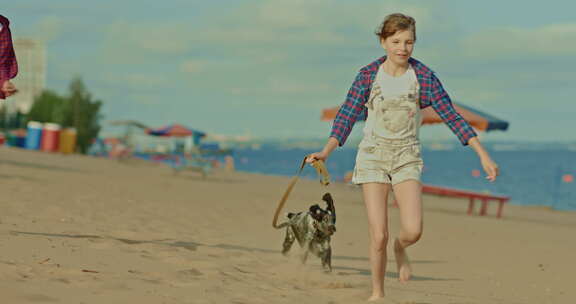 有趣的女孩用皮带拴着一只小狗，他们跑过沙滩