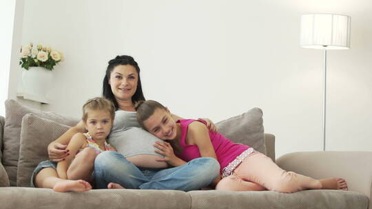 怀孕的女人和她的两个女儿坐在沙发上对着镜头微笑