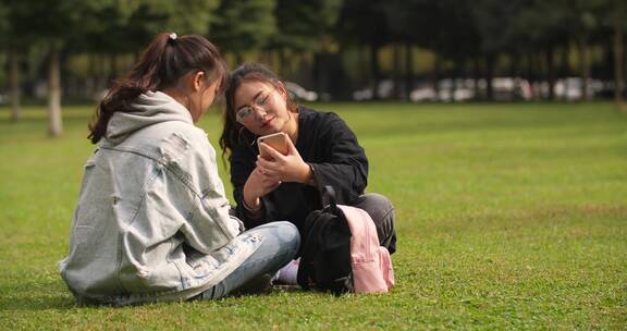 两位女大学生坐在校园草地上看手机学习交谈