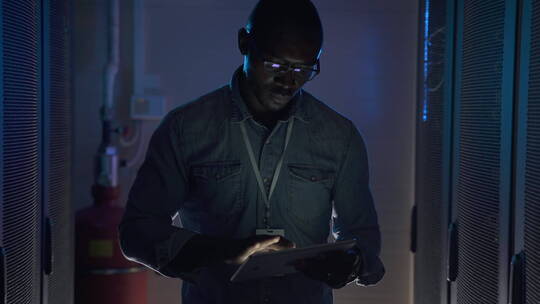 非裔美国信息技术工程师戴着眼镜在半黑暗的服务器机房特写镜头中用平板电脑行走