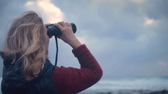 暴风雨天气中，女人站在海边，用双筒望远镜