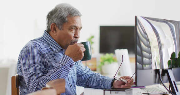 老年混血儿坐在办公桌前喝咖啡，在家使用电脑的视频