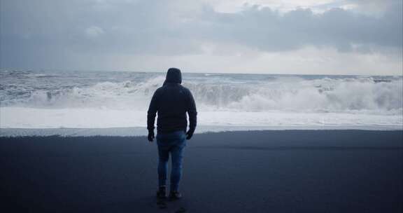 海浪拍打时，穿着夹克的男人站在黑色海滩上