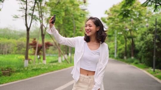 美女走在公园道路上拿着手机开心自拍