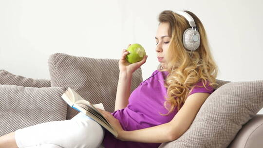 漂亮的年轻女人戴着耳机吃着苹果躺在沙发上看书