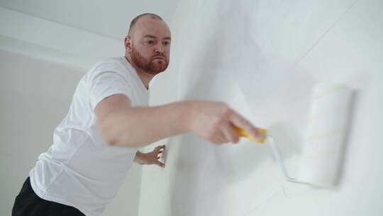 男人用滚筒粉刷白墙。内部维修。年轻的男性装饰师粉刷墙壁视频素材模板下载