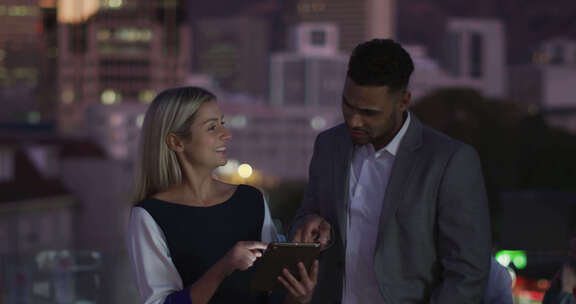两个快乐的不同男性和女性同事晚上在办公室交谈和使用平板电脑的视频