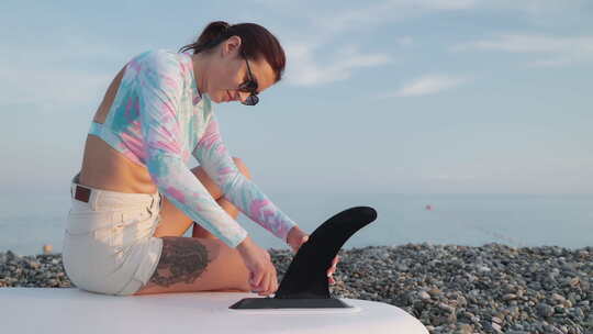 女子在海滩上的SUP板上安装鳍为SUP冲浪和SUP旅游准备设备