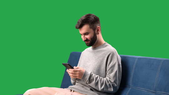 微笑的男人浏览互联网聊天社交网络沙发上的智能手机色度键绿屏