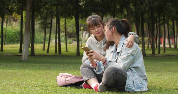 两位女大学生坐在校园草地上看手机学习交谈