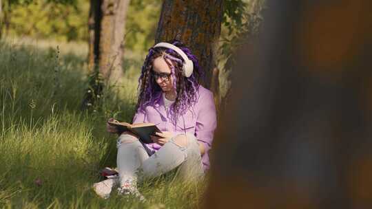 戴眼镜耳机的年轻女子在夏天坐在树旁看书