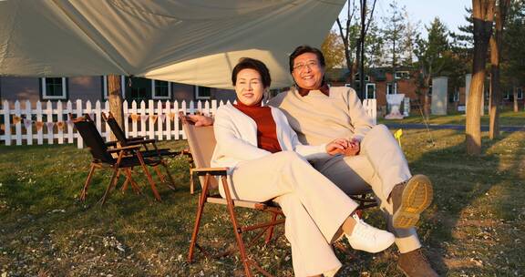 中老年夫妻搂着肩膀坐在露营地天幕下看夕阳