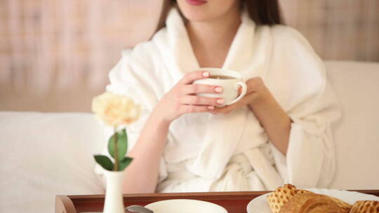 穿着浴袍的女孩坐在床上喝茶看着镜头微笑视频素材模板下载
