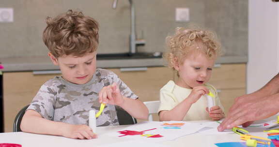 小女孩和小男孩正在用彩纸和胶水和他们的父亲做应用