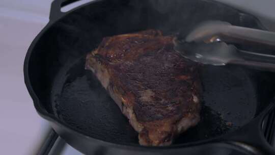 晚餐牛排在铸铁煎锅上烹饪，准备用不锈钢钳子翻转