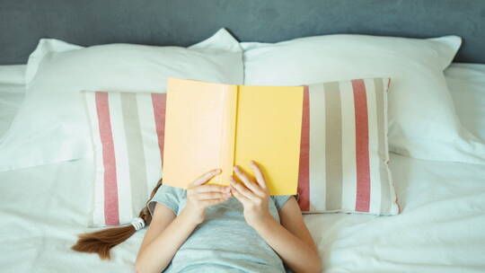 女孩躺在床上看书并对着镜头微笑