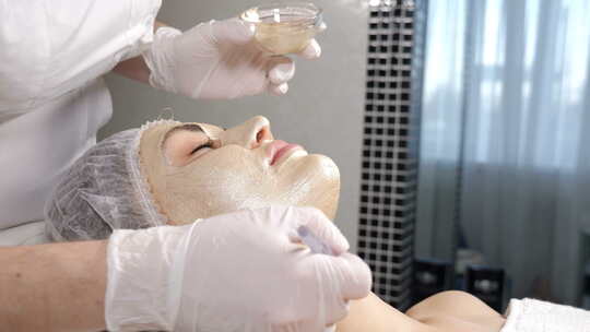 美容概念。美容师在美丽的女性面部皮肤上涂抹化妆品金色面膜。面膜