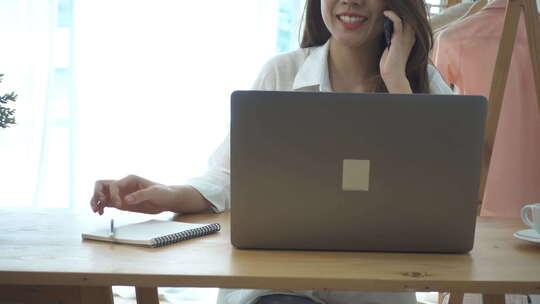 年轻微笑的亚洲女人坐在家里的客厅里打电话，一边用笔记本电脑工作。
