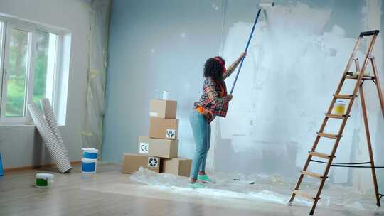 戴红色耳机的非裔美国妇女使用长油漆滚筒用白色油漆粉刷墙壁视频素材模板下载