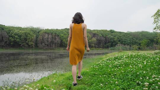 清晨苗条女孩穿橙色长裙走在湖边野花草地
