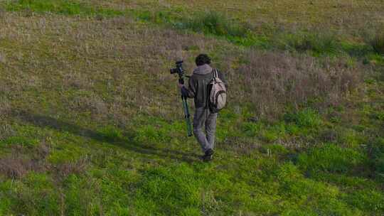 男摄影师在湖边的田野上用三脚架携带相机。