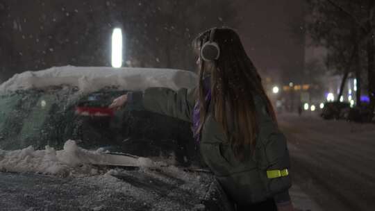 女孩帽子听音乐，晚上用刷子从车里除雪