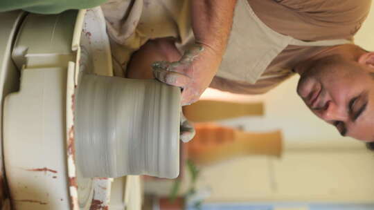 垂直男性陶工穿着围裙坐在车间转盘塑造粘土