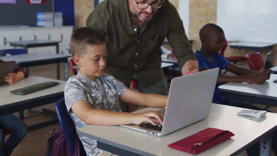 多样化的男老师帮助一个坐在教室里的男生使用笔记本电脑