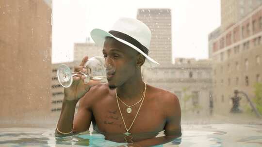 戴着软呢帽和金项链首饰的男人在屋顶游泳池里的玻璃杯里喝酒。词