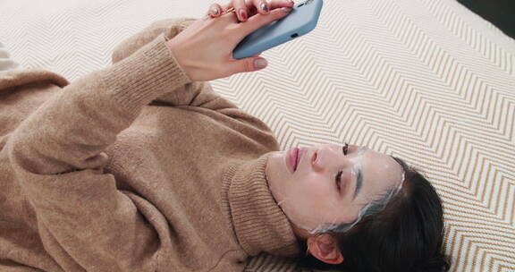 年轻女孩躺在床上看手机敷面膜