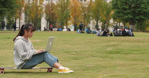 秋天校园女大学生坐在草地滑板上用电脑学习