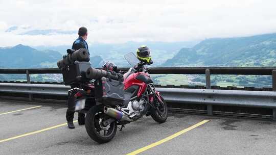 骑着带行李袋的旅游摩托车的自行车手站在山景阿尔卑斯山旁